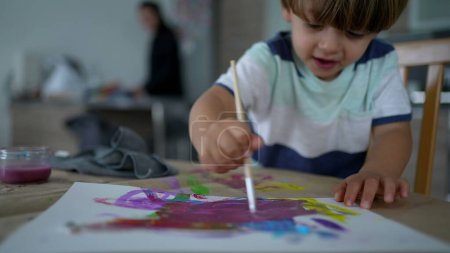 Foto de Niño artístico jugando con la pintura de acuarela - Pintura creativa del muchacho como madre hace las tareas en el fondo - Imagen libre de derechos