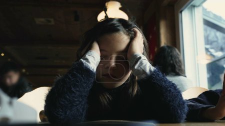 Müdes, gelangweiltes kleines Mädchen bedeckt beim Abendessen im Restaurant das Gesicht mit den Händen, trauriges Kind fühlt sich erschöpft und gelangweilt