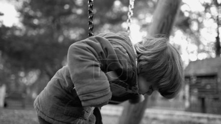 Téléchargez les photos : Enfant seul déprimé s'appuyant sur la balançoire du parc se sentant triste et sombre regardant vers le bas à l'extérieur. Un petit garçon aux prises avec une maladie mentale en monochromatique, noir et blanc - en image libre de droit