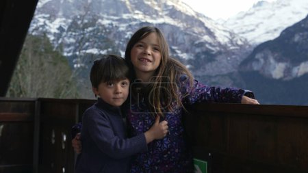 Photo for Young Siblings posing for photo standing in chalet balcony with mountains in background. Little brother and sister embrace during family vacation in the Alps - Royalty Free Image