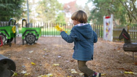 Foto de Un niño pequeño recogiendo hojas amarillas del parque público durante la temporada de otoño. Niño con chaqueta azul juega y corre con follaje en la mano durante el día de otoño - Imagen libre de derechos