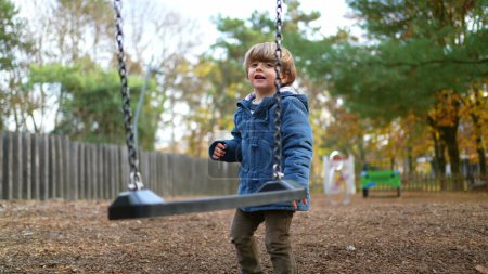 Photo for Child engaged in play outside at park swing, twisting and turning in carefree manner during solo play amidst orange autumn day foliage. Happy Little boy leaning on swing - Royalty Free Image