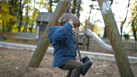 Foto de Niño intenta sentarse en el columpio del parque fuera con chaqueta azul durante el día de otoño - prueba de la infancia y el concepto de error - Imagen libre de derechos