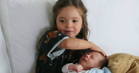 Téléchargez les photos : Sister holding newborn baby infant kissing showing love and affection to brother asleep - en image libre de droit