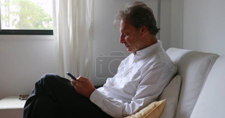 Téléchargez les photos : Candid senior man seated at home sofa checking smartphone working home office - en image libre de droit