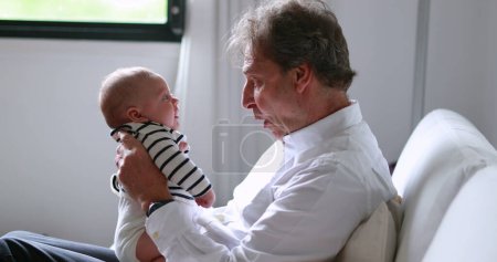 Téléchargez les photos : Candid Grand-père tenant bébé nouveau-né petit-fils dans le canapé intérieur authentique interaction grand-père - en image libre de droit