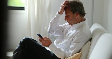 Téléchargez les photos : Candid older man looking at smartphone device at home sofa - en image libre de droit