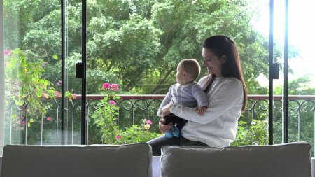 Téléchargez les photos : Moment familial décontracté maman tenant bébé fils bébé à côté de la fenêtre de la maison - en image libre de droit