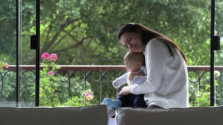 Téléchargez les photos : Candid mère étreignant bébé fils attentionné et aimant à côté de la fenêtre de la maison - en image libre de droit