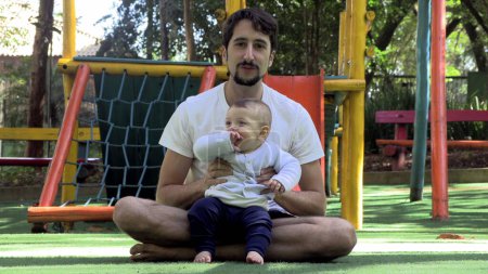 Téléchargez les photos : Père et bébé fils ensemble au parc papa agitant les bras de bébé en disant bonjour à la caméra - en image libre de droit
