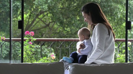 Téléchargez les photos : Candid mère étreignant bébé fils attentionné et aimant à côté de la fenêtre de la maison - en image libre de droit