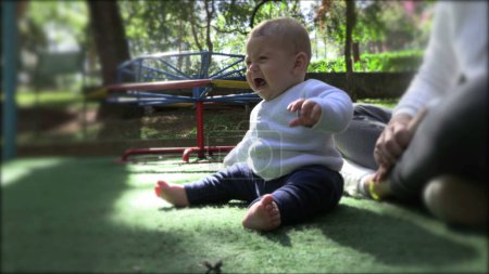 Téléchargez les photos : Bébé tout-petit bouleversé assis au parc de l'aire de jeux à l'extérieur - en image libre de droit