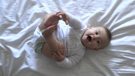 Téléchargez les photos : Bébé garçon mignon au lit regardant la caméra souriant et tenant les pieds - en image libre de droit