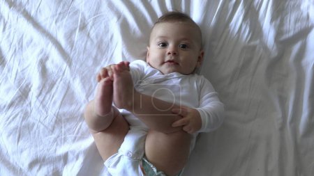 Photo for Cute baby boy in bed looking to camera smiling and holding feet - Royalty Free Image