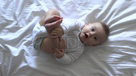 Téléchargez les photos : Bébé garçon mignon au lit regardant la caméra souriant et tenant les pieds - en image libre de droit