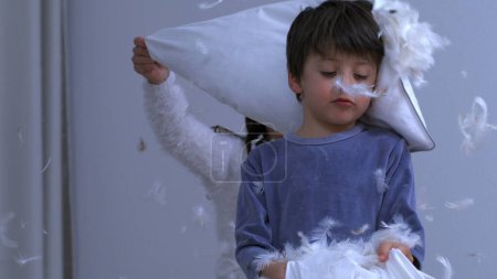 Téléchargez les photos : Enfants qui s'amusent au milieu d'une bataille d'oreillers, petit frère couvert de plumes pendant le jeu - en image libre de droit