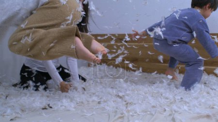 Photo for Children having fun covered in feathers bouncing in bed capturing a happy family moment in super slow motion at 1000 fps. Daughters jumping in bed while mom throws plumage in the air - Royalty Free Image