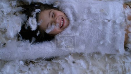 Carefree joyful child covered with feathers amidst happy childhood moment captured in slow-motion. Little girl feeling free laid on plumage surface