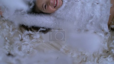Happy Little Girl Falling on bed with thousands of feathers flying in the air, captured with a High-Speed Camera at 1000 fps. Child smiling amidst plumage