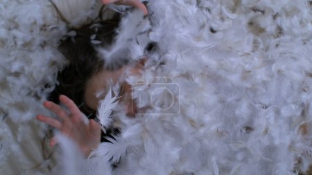 Petite fille allongée dans un lit de plumes, insouciante et joyeuse. plumage tombant au ralenti à 1000 ips couvrant l'enfant de plumes