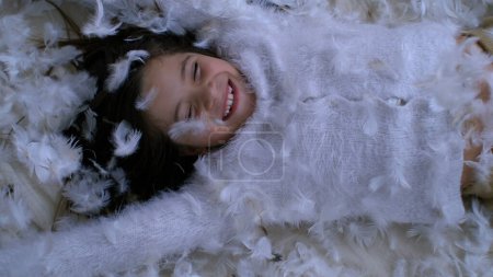 Carefree joyful child covered with feathers amidst happy childhood moment captured in slow-motion. Little girl feeling free laid on plumage surface