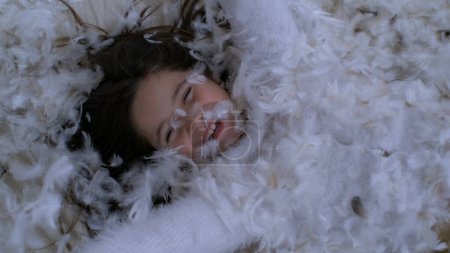 Petite fille allongée dans un lit de plumes, insouciante et joyeuse. plumage tombant au ralenti à 1000 ips couvrant l'enfant de plumes
