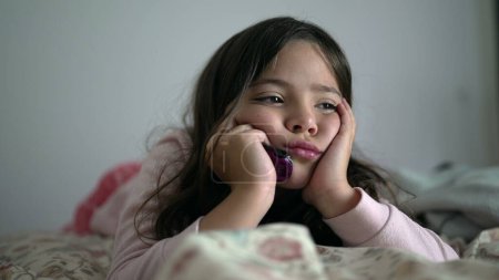 Photo for One contemplative young girl with sad thoughtful expression feeling slight boredom while holding head with hands in chin, 8 year old child laid in bed thinking with nothing to do - Royalty Free Image