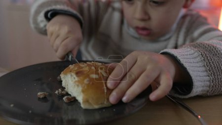 Young Boy schneidet gekonnt Brot für Snacks und die Zubereitung von Kohlenhydraten und nimmt einen Bissen kohlenhydratreiche Nahrung zu sich. 5-jähriges Kind nascht