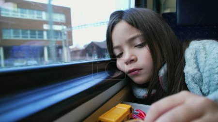 Foto de Una niña aburrida viajando en tren juega con coches de juguete para pasar el tiempo luchando con el aburrimiento. Juego en solitario de niña de 8 años con la cara apoyada en la ventana - Imagen libre de derechos