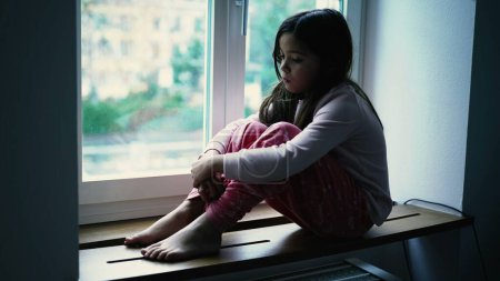 Téléchargez les photos : Petite fille triste mélancolique assise près de la fenêtre de l'appartement, regardant la vue dans une contemplation tranquille. Enfant réfléchi dans une introspection mentale profonde, dépeignant la solitude de l'enfance - en image libre de droit