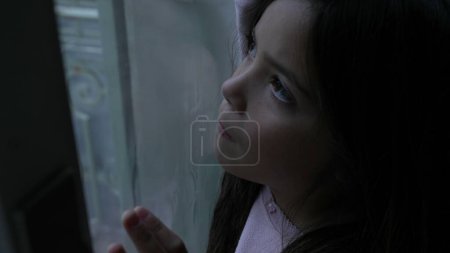 One pensive little girl leaning on glass window in deep mental introspection. Thoughtful childhood contemplation, child struggling with solitude at home