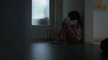 Téléchargez les photos : Triste enfant solitaire assis dans le couloir de la maison, dans une sombre scène maussade couvrant le visage d'anxiété et de dépression. Enfant aux prises avec une maladie mentale dans l'enfance - en image libre de droit