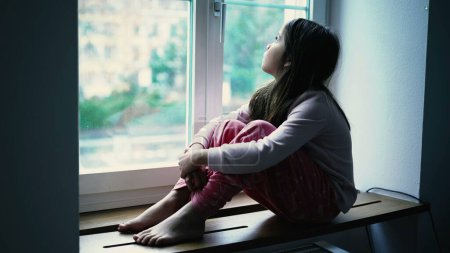 Foto de Niña triste melancólica sentada junto a la ventana del apartamento mirando a la vista en contemplación tranquila. Niño reflexivo en profunda introspección mental, que representa la soledad infantil - Imagen libre de derechos