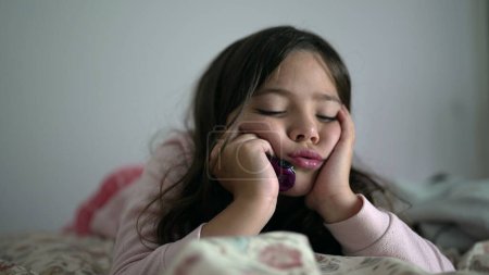 Photo for One contemplative young girl with sad thoughtful expression feeling slight boredom while holding head with hands in chin, 8 year old child laid in bed thinking with nothing to do - Royalty Free Image