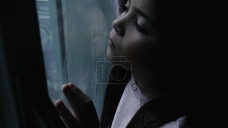 Foto de Niño solitario sintiéndose triste y deprimido en la escena de invierno malhumorado por la ventana de cristal. Una niñita pensativa atrapada en casa - Imagen libre de derechos