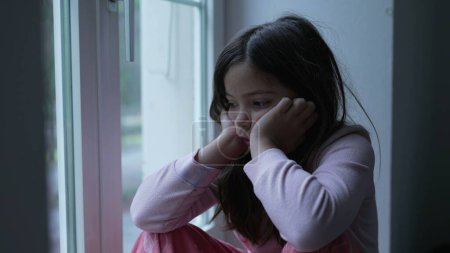 Foto de Triste niña pensativa sintiendo soledad por ventana del apartamento mirando a la vista con mirada reflexiva. Niño deprimido contemplativo - Imagen libre de derechos