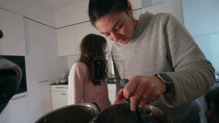 Mother preparing food next to her daughter while son roams around the kitchen, authentic family lifestyle scene parent cooking holding spatula in hand mixing ingredients