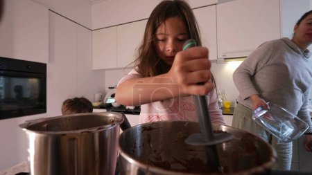 Téléchargez les photos : Petite fille concentrée remuant pot rempli de chocolat, préparer le gâteau avec la mère et frère debout à la cuisine. Parent et enfants cuisinent ensemble, authentique moment de style de vie familial - en image libre de droit