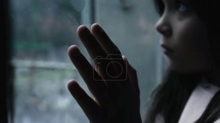 Childhood mental illness depiction of little girl struggling with depression and worthlesness leaning on glass window staring at view in moody scene