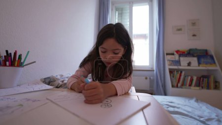 Téléchargez les photos : Petite fille concentrée dessinant sur papier dans la chambre à coucher, enfant utilisant un stylo. Développement artistique de l'enfance, temps libre - en image libre de droit