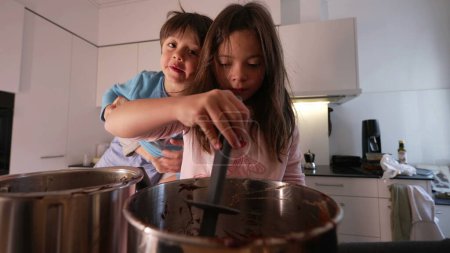 Photo for Concentrated Little girl stirring pot filled with chocolate, preparing cake with little brother standing at kitchen. Children cooking together, authentic family lifestyle moment - Royalty Free Image