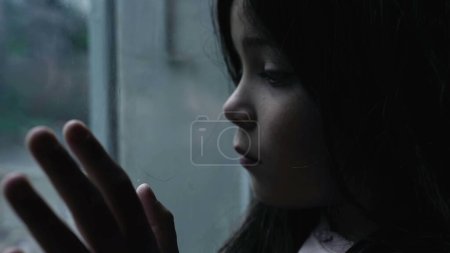 Childhood mental illness depiction of little girl struggling with depression and worthlesness leaning on glass window staring at view in moody scene
