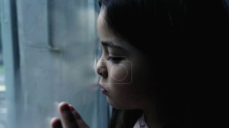 Foto de Un niño deprimido apoyado en una ventana de cristal sintiéndose triste y solo en casa, una pequeña niña introspectiva depicitng enfermedad mental infantil - Imagen libre de derechos