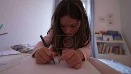Téléchargez les photos : Une petite fille concentrée dessinant sur du papier dans sa chambre. L'enfant dessine avec un stylo. Enfance loisirs artistiques artisanat temps - en image libre de droit