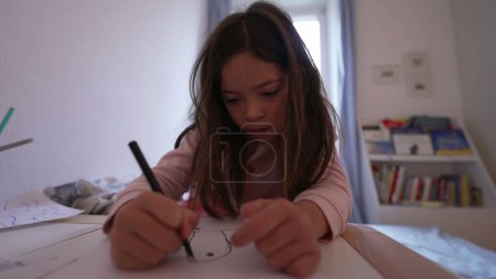 Téléchargez les photos : Une petite fille concentrée dessinant sur du papier dans sa chambre. L'enfant dessine avec un stylo. Enfance loisirs artistiques artisanat temps - en image libre de droit