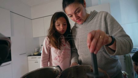 Mother preparing food next to her daughter while son roams around the kitchen, authentic family lifestyle scene parent cooking holding spatula in hand mixing ingredients