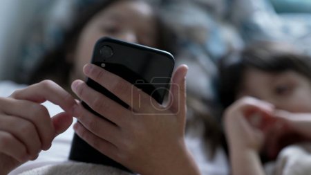 Photo for Children looking at phone screen - sister holding cellphone while small brother stares at media online, kids lying in bed under bedsheets engaged with modern technology - Royalty Free Image
