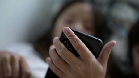 Photo for Little girl holding smartphone device looking at screen consuming media entertainment online. 8 year old child engaged with modern technology lying in bed hypnotized by phone - Royalty Free Image
