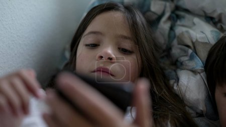 Foto de Niña sosteniendo dispositivo de teléfono inteligente mirando la pantalla que consume entretenimiento multimedia en línea. Niño de 8 años comprometido con la tecnología moderna acostado en la cama hipnotizado por teléfono - Imagen libre de derechos