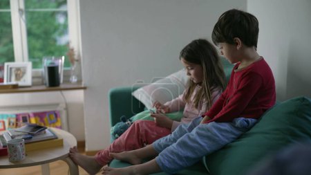 Photo for Little brother and sister looking at cellphone screen while seated on couch sofa in living room. Children hypnotized by content online sharing gadget - Royalty Free Image